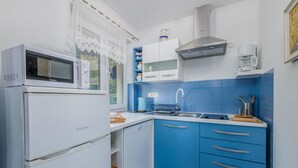 A compact kitchen with white appliances, blue cabinets, and a window with lace curtains. - 1 bedroom awesome apartment in Stara Baska (Stara Baska)