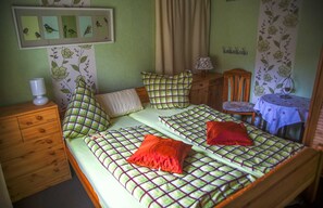A bedroom with two single beds, a wooden dresser, a small table with a lamp, and a wall with bird and floral wallpaper.