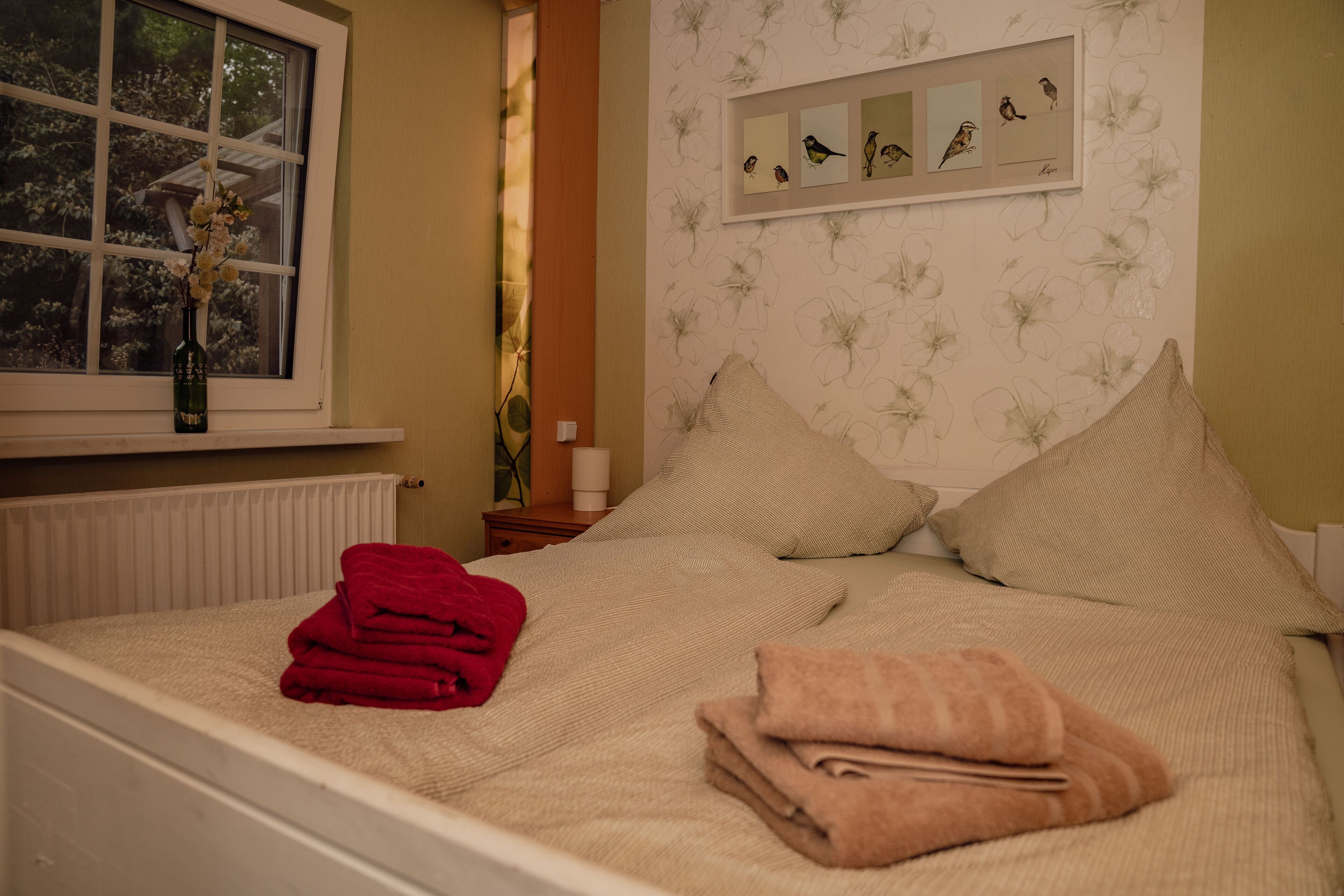 A bedroom with a bed, pillows, a window with a vase, and a wall with bird illustrations.