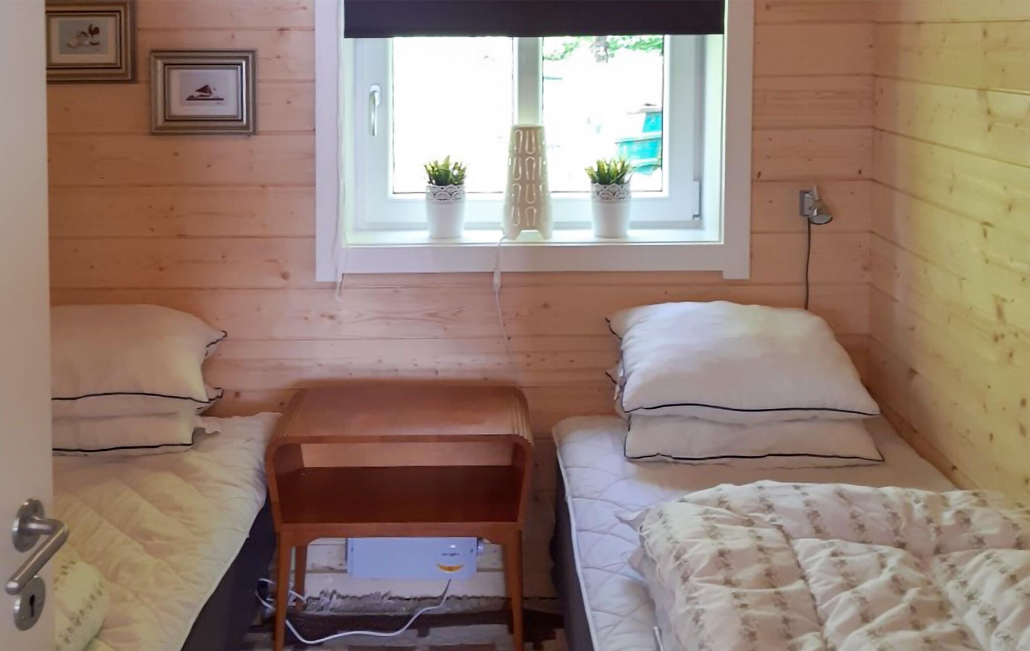A small, cozy room with two beds, a window with potted plants, and a wooden wall.