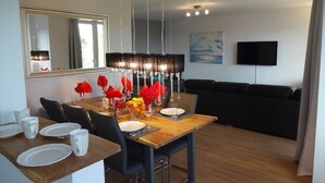 Un comedor moderno con una mesa de madera dispuesta para cuatro personas, un sofá negro, un televisor de pantalla plana montado en la pared y un espejo grande sobre la mesa.