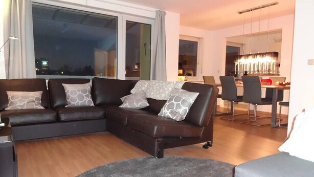 Una sala de estar moderna con un sofá seccional, un comedor con sillas y un amplio ventanal que ofrece vistas de la ciudad de noche.