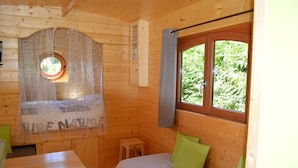 A wooden cabin room with a bed, a small table, a window with curtains, and a view of greenery outside. - Unusual gite equipped to receive up to 5 people (Seux)