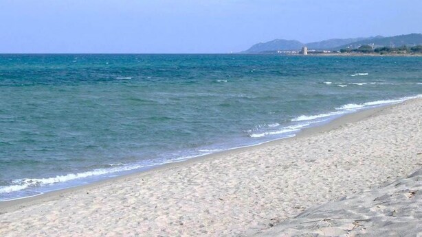 Una playa con agua azul cristalina, orilla arenosa y edificios lejanos sobre un terreno montañoso.