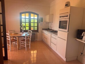 Uma cozinha com mesa de jantar, cadeiras, fogão, geladeira e janela com vista para áreas verdes.
