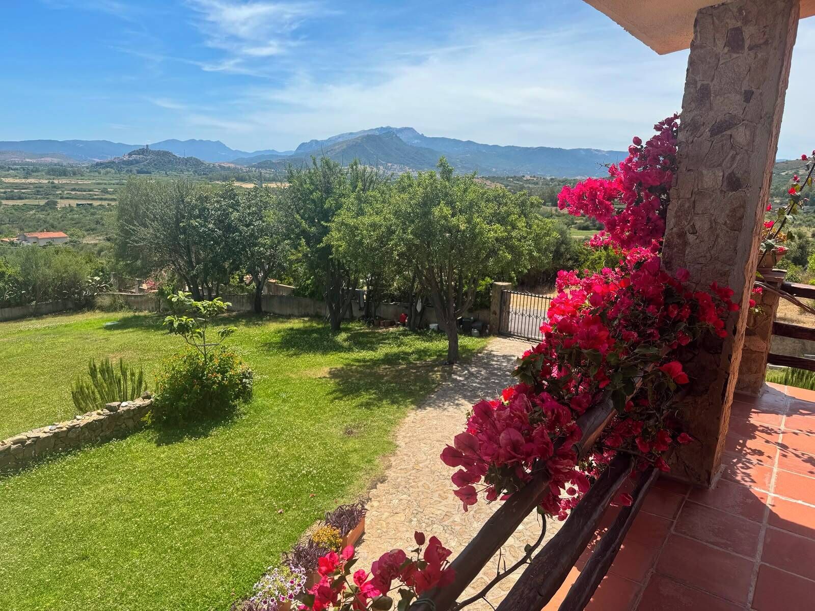 A balcony with a stone pillar, a view of a green landscape, and a mountain range in the distance.
