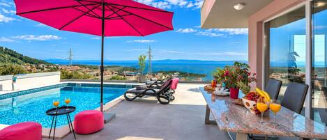 A poolside area with a red umbrella, lounge chairs, and a dining table with fruit and drinks.