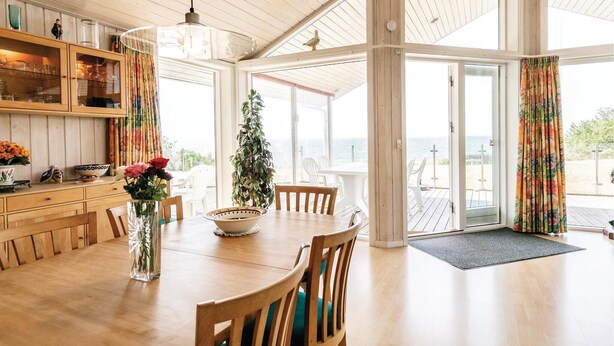 A spacious dining area with a wooden table and chairs, a vase with flowers, and a view of the sea through large glass doors. - 4 bedroom awesome home in Rønne (Rønne)