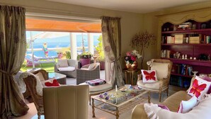 A well-furnished living room with a view of the sea, a bookshelf, and a coffee table with books and decorative items.