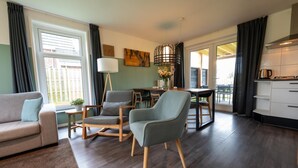 Ein modernes Wohnzimmer mit einer Couch, zwei Sesseln, einem Esstisch mit Stühlen, einer Lampe und einer Küchenzeile mit Schränken.