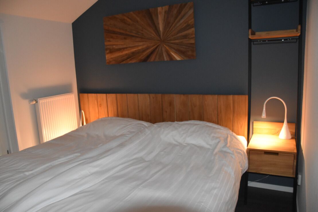 A bedroom with a wooden headboard, a white bedspread, a bedside table with a lamp, and a wall-mounted shelf.