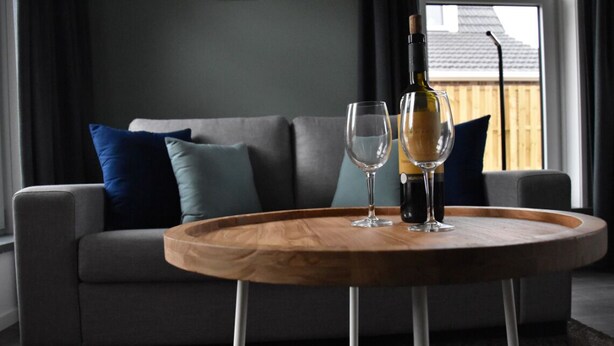 Ein modernes Wohnzimmer mit grauem Sofa, blauen und türkisen Kissen, einem Holztisch mit einer Weinflasche und zwei Gläsern sowie Blick auf eine Holzterrasse durch ein Fenster.