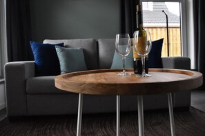 Ein modernes Wohnzimmer mit grauem Sofa, blauen und türkisen Kissen, einem Holztisch mit einer Weinflasche und zwei Gläsern sowie Blick auf eine Holzterrasse durch ein Fenster.