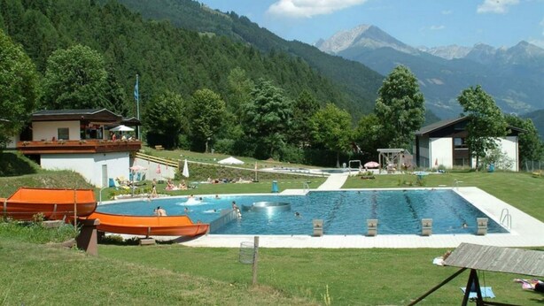 Piscina com vista para montanhas, cercada por edifícios e uma área ajardinada.