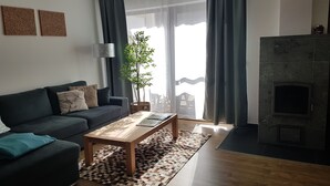 Uma sala de estar com um sofá escuro, uma mesa de centro de madeira e um tapete estampado. Há uma lareira e uma janela grande com cortinas.