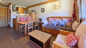 A cozy living area with a wooden dining table, chairs, and a sofa with patterned cushions. A clock and a framed picture adorn the wall. - Cozy apartment at the foot of the slopes, pets allowed (Les Menuires)