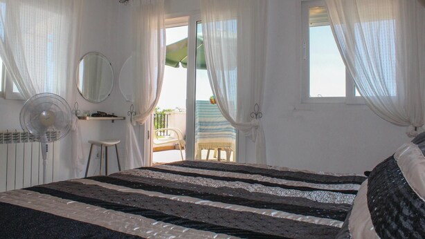 Una camera da letto con un letto, una ventola, una sedia, un tavolino, uno specchio e un balcone con una sedia e un asciugamano a righe.
