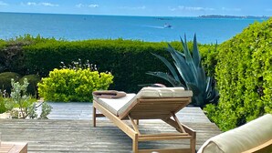 Une terrasse en bois avec deux chaises longues et une petite table, d’où l’on peut admirer un plan d’eau avec des bateaux et une côte lointaine.