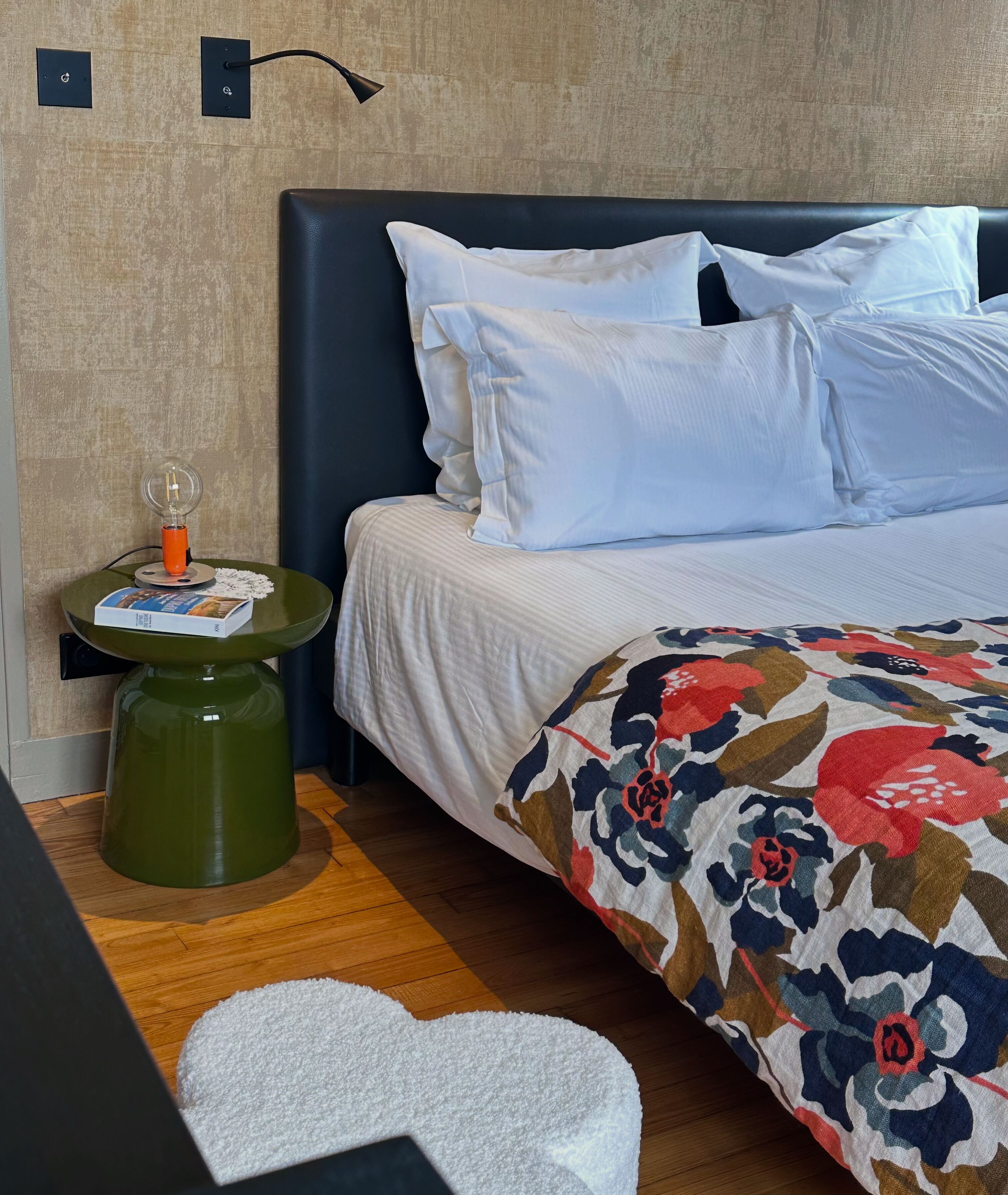 Ein ordentlich bezogenes Bett mit weißen Leinen und einer floralen Decke, ein kleiner Nachttisch in Grün mit einer Lampe und einem Buch darauf.