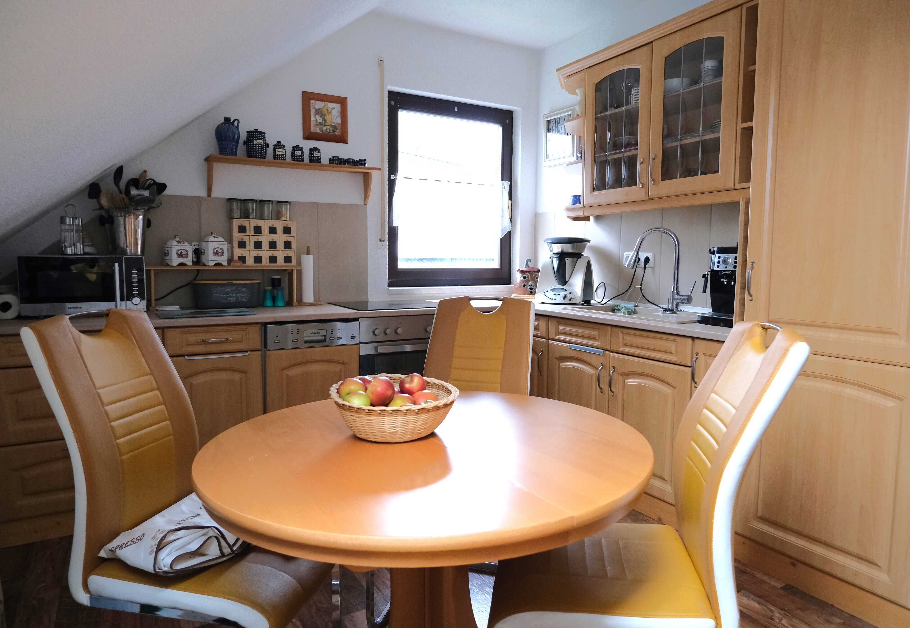 A kitchen with wooden cabinets, a dining table with chairs, a microwave, and a window with blinds.