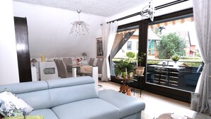A modern living room with a light blue sofa, a dining area with a table and chairs, and a balcony with potted plants and a view of the neighborhood. - Apartment with balcony in a quiet, sunny location on the outskirts of Saxon Switzerland (Sebnitz)