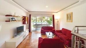 Un salón moderno con televisor de pantalla plana, sofás rojos, una mesa de centro de vidrio y vistas a una piscina y jardín a través de grandes puertas correderas de cristal.