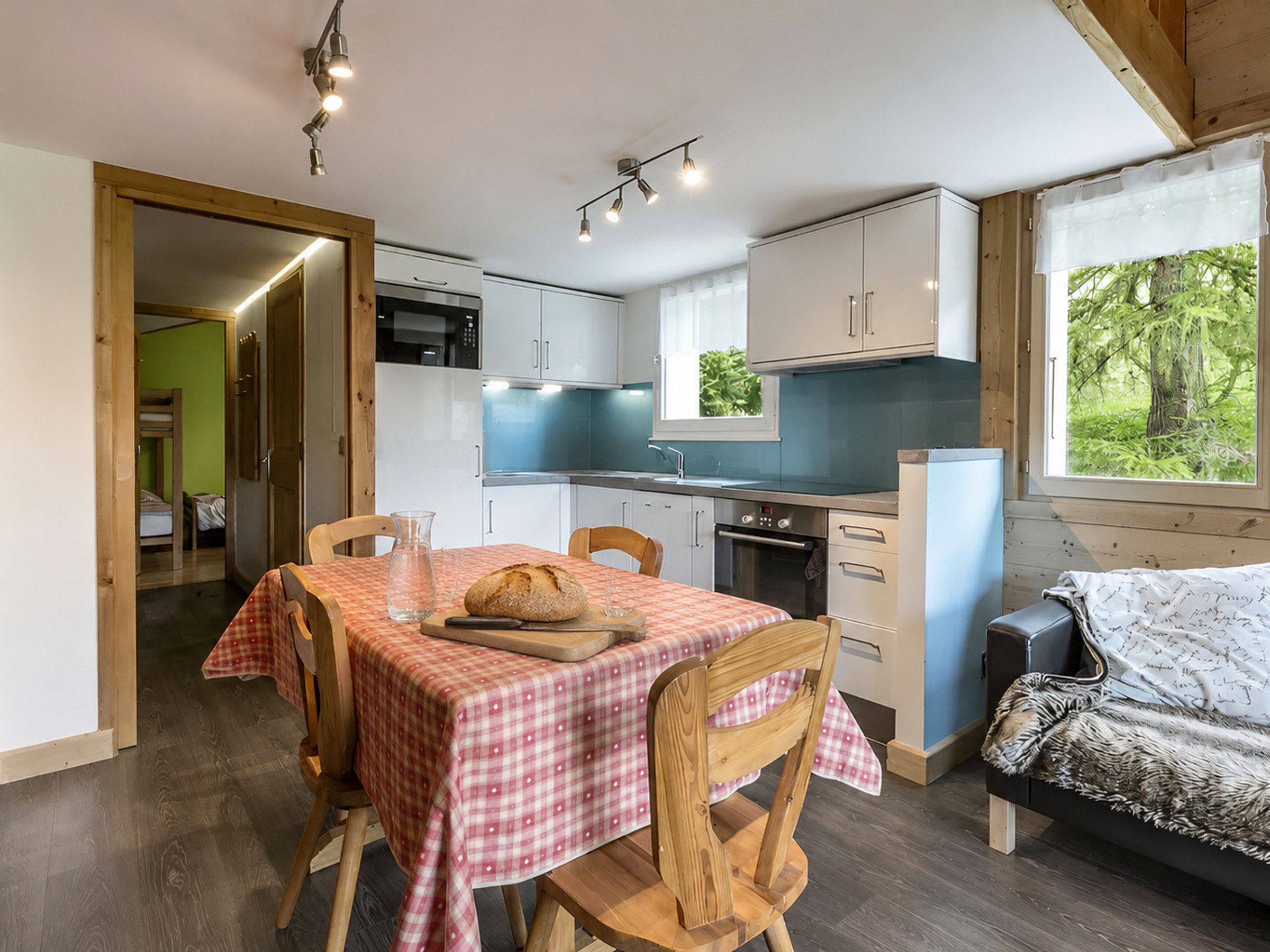 Une cuisine compacte dotée d’appareils électroménagers modernes, un coin repas avec une nappe à carreaux et un espace nuit confortable offrant une vue sur la verdure.