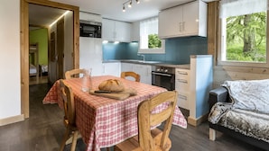 A compact kitchen with modern appliances, a dining area with a checkered tablecloth, and a cozy sleeping area with a view of greenery. - Charming 3* apartment, near slopes, wood stove, garage, WIFI (La Clusaz)