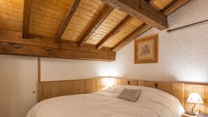A bedroom with a wooden ceiling, a bed with white bedding, a nightstand with a lamp, and a framed picture on the wall. - Charming 3* apartment, near slopes, wood stove, garage, WIFI (La Clusaz)