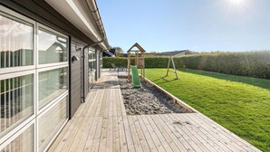 Una casa moderna con ventanas amplias, una terraza de madera, una zona de juegos con tobogán y un césped cuidado.