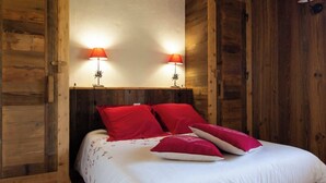 Quarto com painéis de madeira, uma cama com roupa de cama vermelha e branca, duas luminárias fixadas na parede e teto de madeira.
