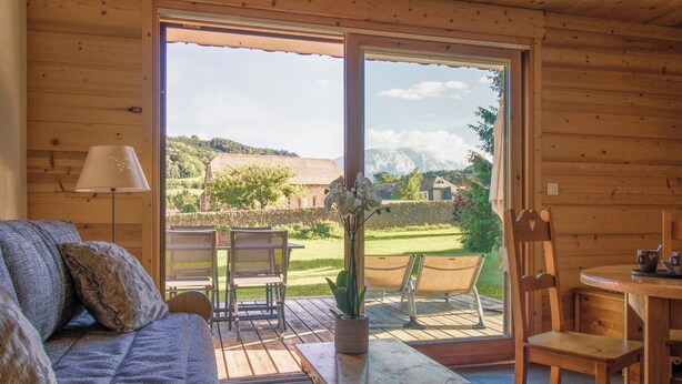 Interior acolhedor em madeira, com sofá, mesa, cadeiras e uma grande porta de vidro deslizante que dá para área externa com vista para paisagem verde e montanhas.