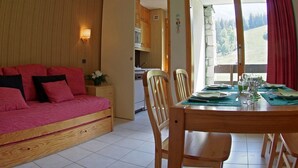 Een gezellige kamer met een houten bank, een eettafel gedekt voor vier personen, en een keuken die zichtbaar is door een open deur.