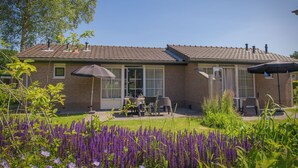 Una casa de ladrillo con techo de tejas, un patio con mobiliario exterior y un jardín con flores moradas.