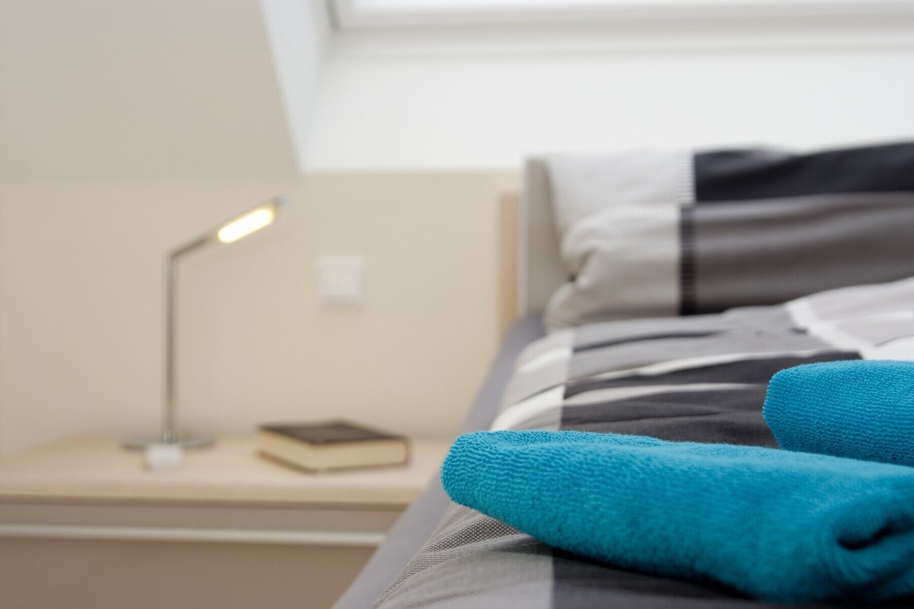 A neatly made bed with a blue towel, a bedside lamp, and a book on a nightstand.