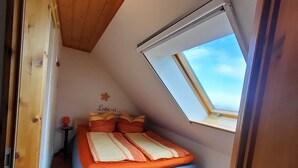 Ein kleines, gemütliches Schlafzimmer mit geneigter Decke, einem Oberlicht, einem Bett mit orangefarbener Bettwäsche und einem kleinen Tisch mit einer Lampe.