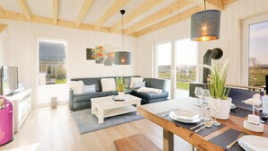 Un salon moderne avec un canapé, une table basse et une table à manger dressée pour un repas. De grandes fenêtres offrent une vue sur l’extérieur.