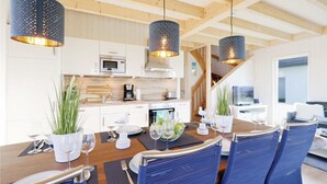 Une cuisine moderne avec des meubles blancs, un four à micro-ondes et une machine à café. On y trouve un coin repas avec une table en bois pour quatre personnes, des chaises bleues et une grande fenêtre.