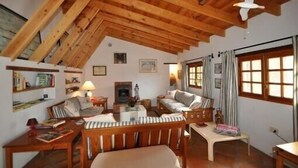 A cozy room with a sloped ceiling, wooden beams, a fireplace, a sofa, a table, and a window with curtains.
