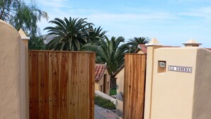 A narrow pathway leading to a wooden gate, with a sign reading "LA GOMERA" on a building to the right.