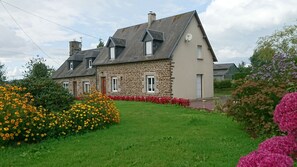 A stone house with a sloped roof, a small attached garage, and a well-maintained lawn with colorful flowers. - country house with free wifi in a quiet area 45 mins from mont st michel (Saint-Laurent-De-Cuves)