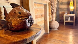 A wooden fish sculpture on a table, a woven basket, a small shelf, and a lit candle in a rustic room with wooden walls and flooring.