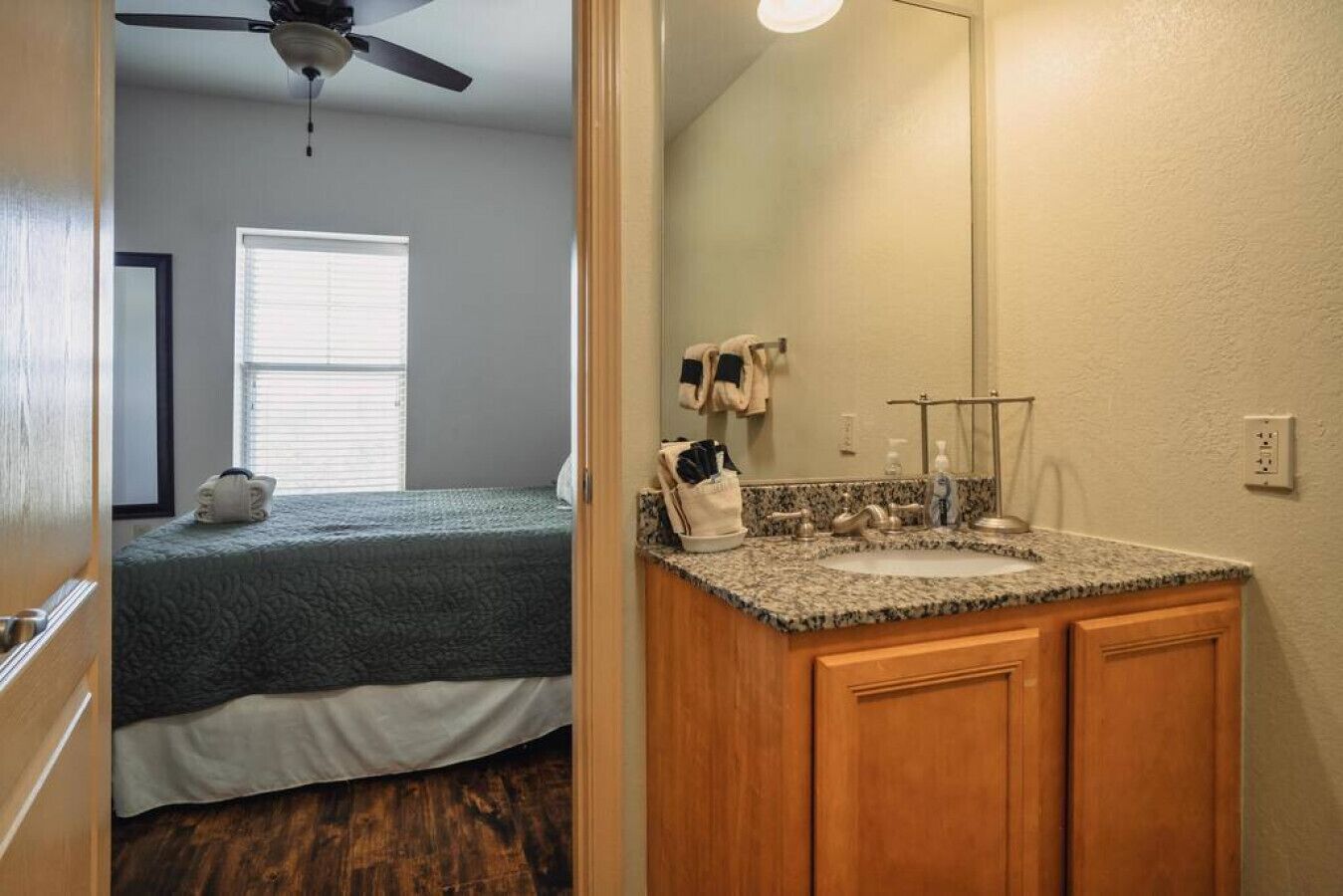 Une chambre à coucher comprenant un lit, un ventilateur de plafond, une fenêtre avec des stores, ainsi qu’une salle de bain avec un lavabo, un miroir et un porte-serviettes.