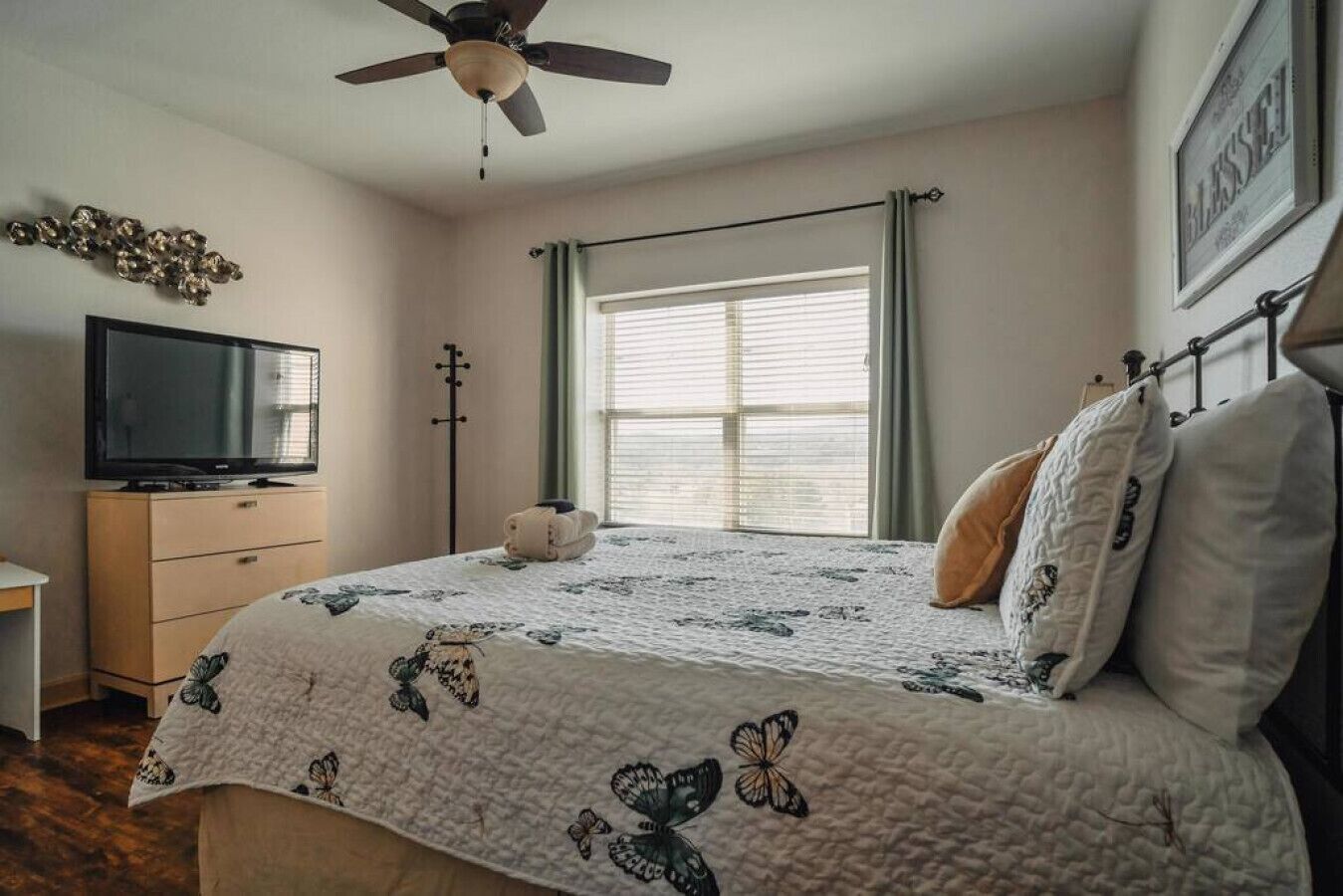 A bedroom with a bed, a television, a ceiling fan, a window with curtains, and a wall-mounted light fixture.
