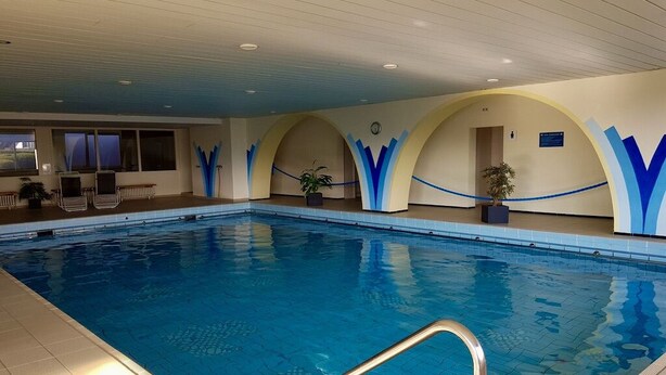 Une piscine intérieure à l’eau d’un bleu limpide, avec un escalier en métal et un espace détente comprenant des chaises et une plante en pot.