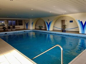 Une piscine intérieure à l’eau d’un bleu limpide, avec un escalier en métal et un espace détente comprenant des chaises et une plante en pot.