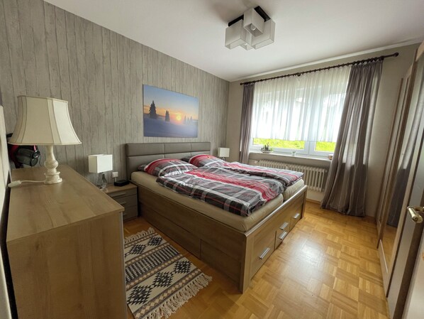 A bedroom with a wooden bed, a bedside table with a lamp, a painting on the wall, and a window with curtains.