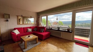 Un salon avec un canapé rouge, une table basse en bois et une vue sur l’extérieur grâce à de grandes fenêtres.