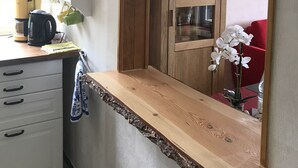 Une cuisine avec des armoires en bois, un plan de travail et un four encastré. Des plantes en pot sont disposées sur le mur, et une chaise rouge se trouve à l’arrière-plan.