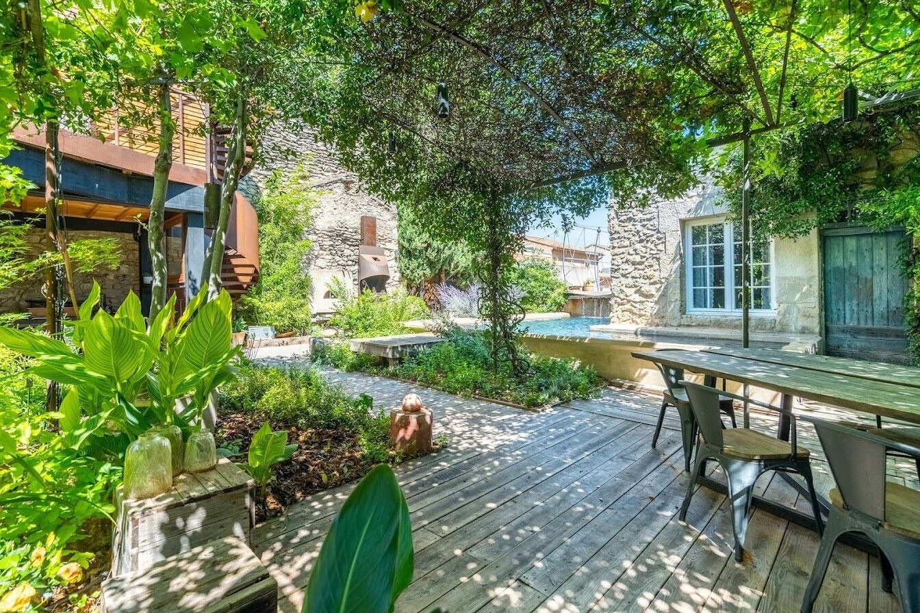Une terrasse en bois avec une table et des chaises, entourée d’une végétation luxuriante et d’un bâtiment en pierre.
